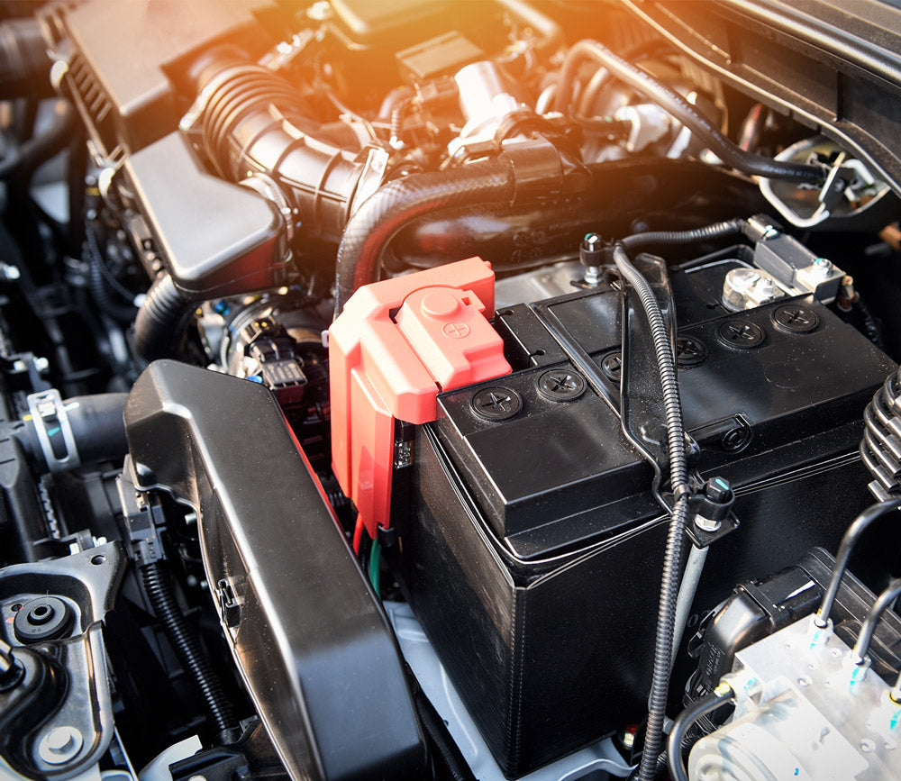 Nahaufnahme eines Automotorraums mit einer schwarzen rechteckigen Batterie, die in der Mitte deutlich zu sehen ist. Die Batterie hat eine rote Schutzabdeckung am Pluspol. Umliegende Komponenten, Kabel und Schläuche sind ordentlich um die Batterie herum angeordnet.