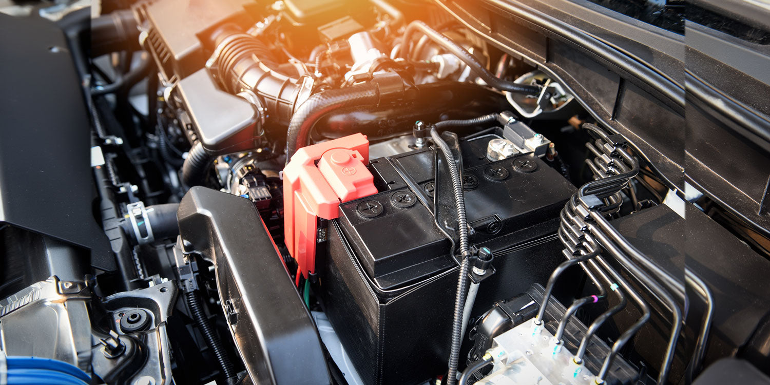 Nahaufnahme eines Automotorraums mit einer prominent platzierten Batterie mit einer roten Schutzabdeckung über einem Anschluss. Verschiedene Motorkomponenten und Kabel sind sichtbar und werden durch helle Lichtverhältnisse hervorgehoben.