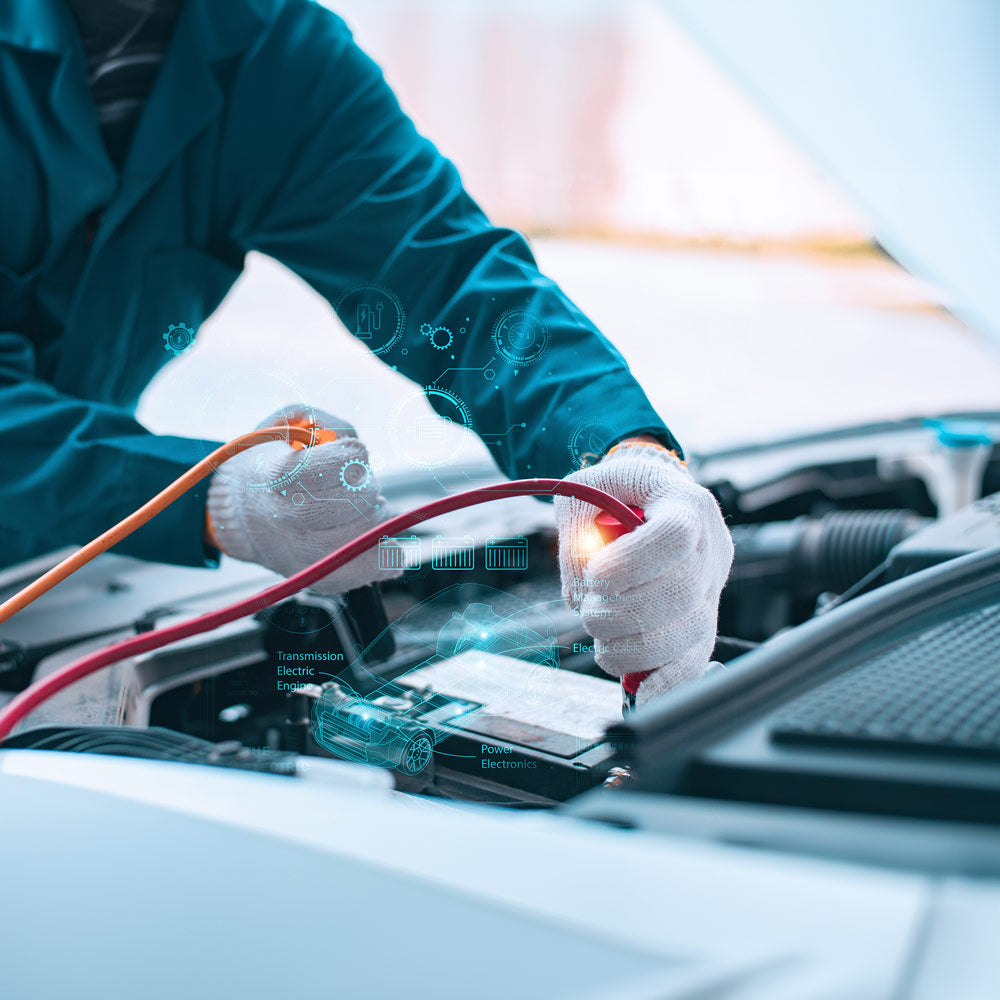 Ein Mechaniker in blaugrüner Jacke und weißen Handschuhen verwendet rote und schwarze Kabel, um eine Autobatterie bei geöffneter Motorhaube anzuschließen. Holografische Symbole und Schemata werden eingeblendet, um auf erweiterte Diagnosefunktionen und Technologieintegration im Fahrzeug hinzuweisen.