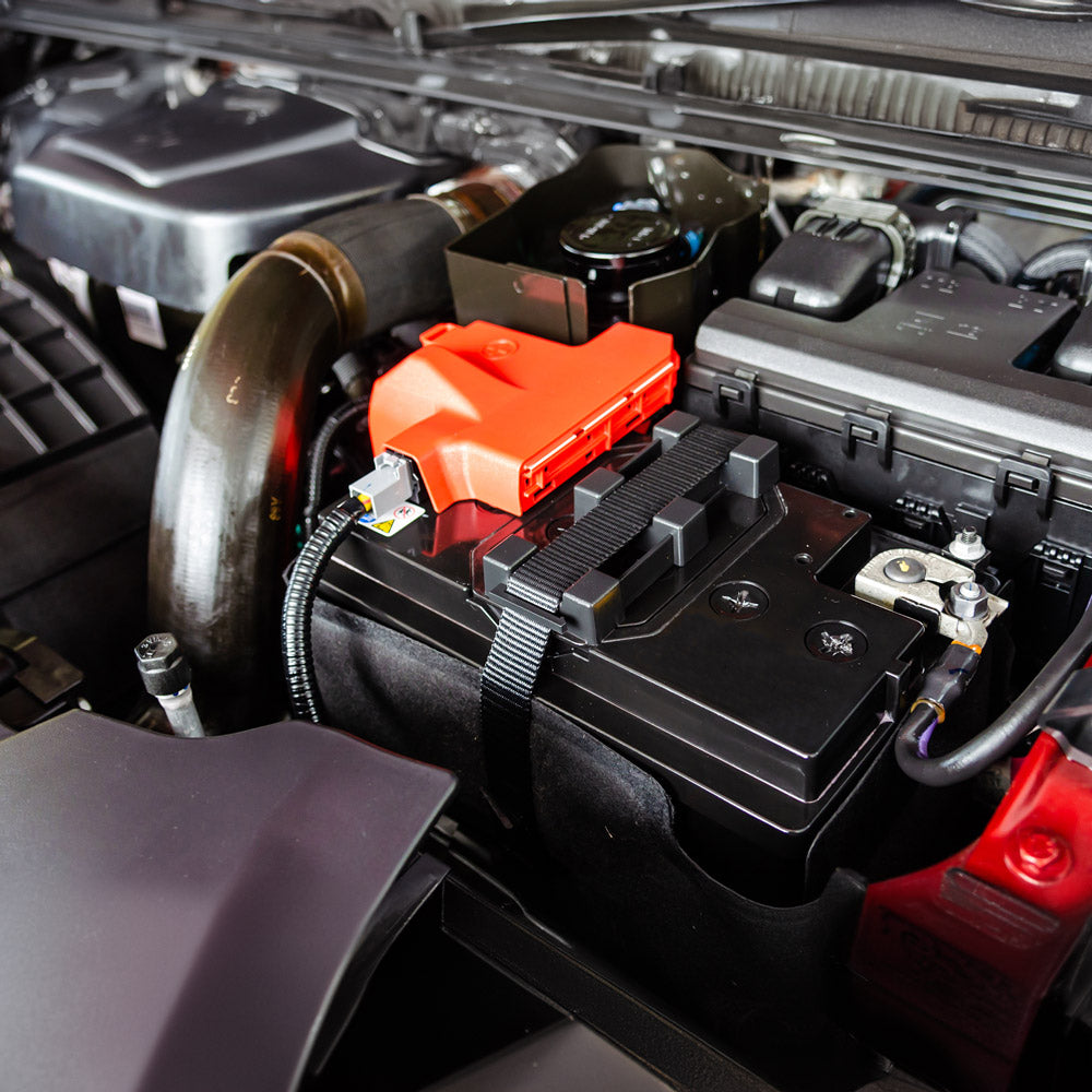 Nahaufnahme des Innenraums eines Automotors mit Fokus auf die Batterie. Die schwarze Batterie hat eine rote Abdeckung über einem Anschluss und ist mit Bändern gesichert. Verschiedene umliegende Motorkomponenten, Kabel und Schutzgehäuse sind ebenfalls sichtbar.