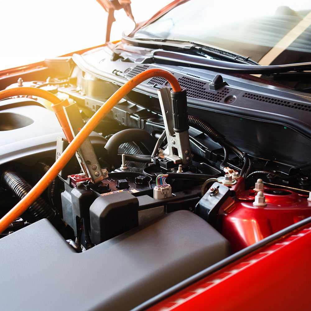 Nahaufnahme eines Automotors, der mit an den Batterieklemmen angeschlossenen Starthilfekabeln überbrückt wird. Die Motorhaube des roten Autos ist geöffnet und Sonnenlicht scheint auf den Motorbereich.