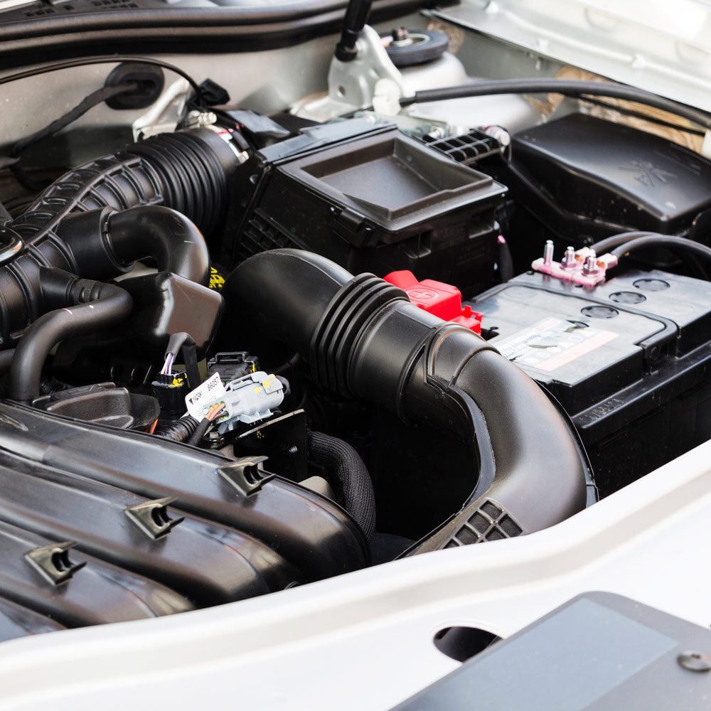 Eine Nahaufnahme des Motorraums eines Autos, in der verschiedene Komponenten wie Batterie, Schläuche, Luftansaugrohre und elektrische Anschlüsse zu sehen sind. Die Teile sind sauber und gut gewartet und im Motorraum des Fahrzeugs untergebracht.